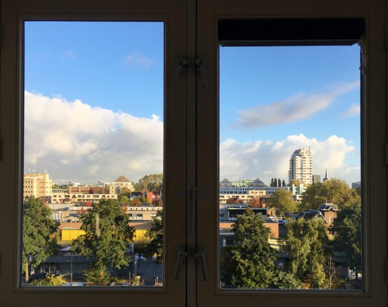 Uitzicht vanuit de noordelijke slaapkamer. Het uitzicht bestaat uit bomen en daarachter en tussen wat daken en gebouwen afsteken tegen een mooie Hollandse lucht. 