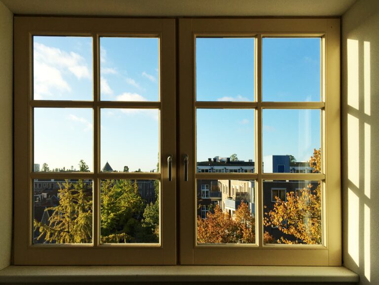 Uitzicht vanuit de zuidelijke slaapkamer. Kijkt uit op een straat met bomen in herfstkleuren en rechts en achter wat gebouwen en blauwe lucht. De ochtendzon schijnt naar binnen
