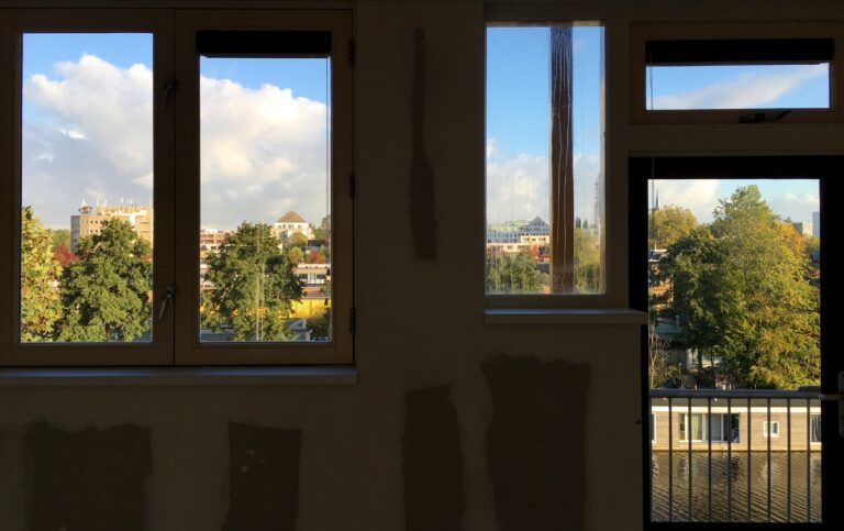 Het uizicht aan de noorzijde van de woonkamer. Bestaande uit twee ramen een tussenraam een deur met glas en een bovenraam. Het is een soort mozaïekje. Je ziet woonboten, bomen en helemaal daarachter wat daken afsteken tegen een mooie Hollandse lucht. 