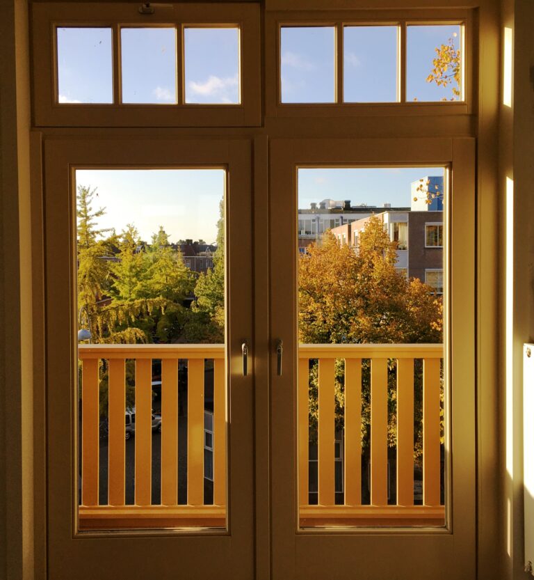 uitzicht vanuit de voordeuren naar het balkon aan de zuidkant van de woonmaker. Kijkt uit op een straat met bomen in herfstkleuren en rechts en achter wat gebuwen en blauwe lucht. De ochtendzon schijnt naar binnen