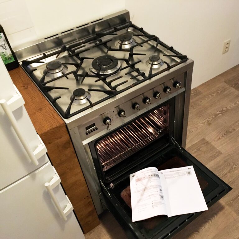 Schuin van boven gefotografeerd stalen Smeg gasfornuis met 5 pitten. De ovendeur staat open en daar ligt de handleiding op.