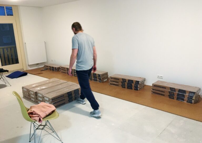 Geschilderde kamer met daarin een aantal stroken kurkvloer. De pakken worden er nu opgelegd zodat we de ruimte hebben om verder te leggen en tevens de gelegde vloer op zijn plaatst te houden.