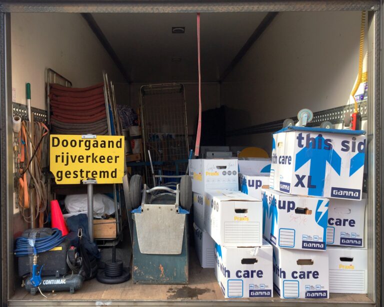 Achterkant van de vrachtwagen met allemaal spullen en dozen erin en een geel bord " Doorgaand rijverkeer gestremd"