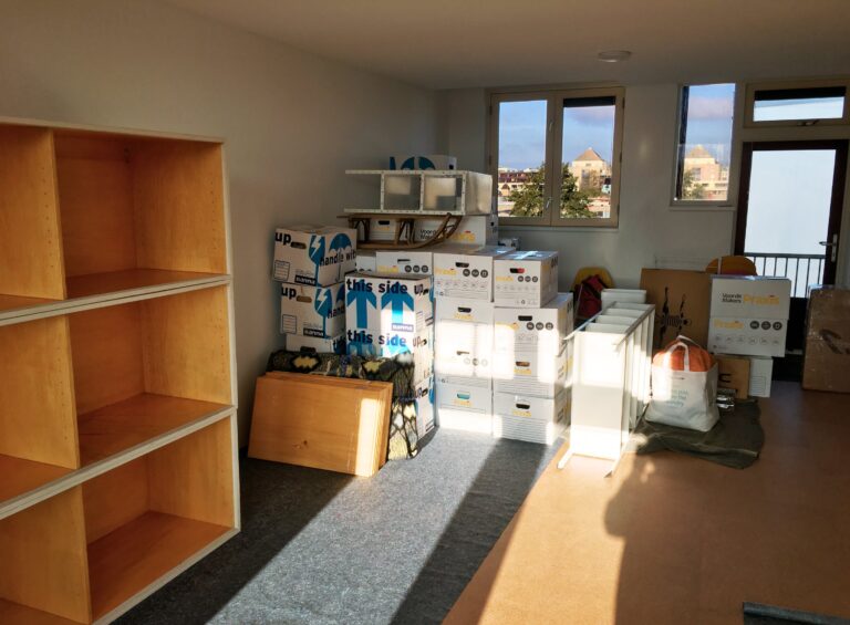 De kamer vol met dozen en een kast met een doorkijkje door de ramen. De zon schijnt heerlijk naar binnen.