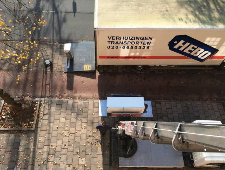 Foto genomen vanaf het balkon. Onder aan de weg staat de verhuiswagen. Een verhuislift heeft een koelkast klaar om naar boven te sturen. 