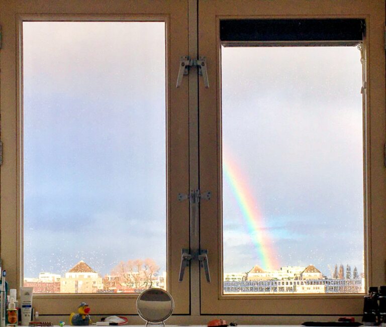 Uitzicht vanuit mijn raam. Bestaat uit twee delen. Het uitzicht is onderaan een streepje gebouwen en verder lucht. En er is in het rechter raam een regenboog te zien.