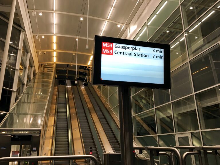 De entreehal van station Ganzenhoef in de avond. Het bestaat helemaal uit glas met wit frame. In het midden een paal met daarop een beeldscherm en de aankomsttijden van de metro's. Erachter de 3 hoge roltrappen naar het perron.
