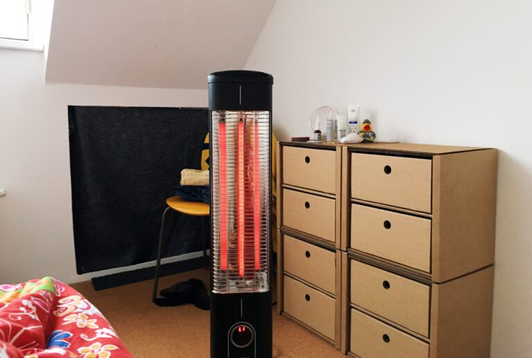 Hoek van een slaapkamer met een schuin dak. Links een ladekast van karton met 8 lades. In het midden een zwarte staande infraroodkachel. Daarachter een gele stoel en linksvoor een stukje dekbed.