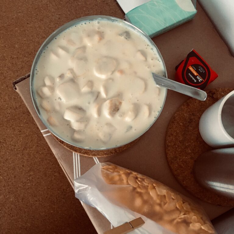 Kak met eten van bovenaf gefotografeerd. Staat op een kartonnen tafel met nog andere attributen zoals twee kurken onderzetters, zakdoekjes, mok, rolmaat, metalen thermosfles en cornflakes.