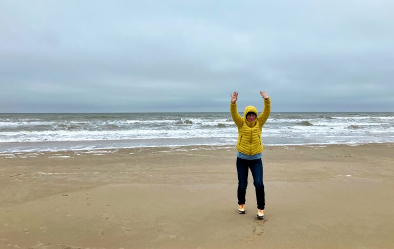 Els met gele jas en donkerblauwe spijkerbroek op het strand. Een stuk Texels stand, de branding en de zee tot aan de horizon. Daarboven een lucht gevuld met grijze wolken.
