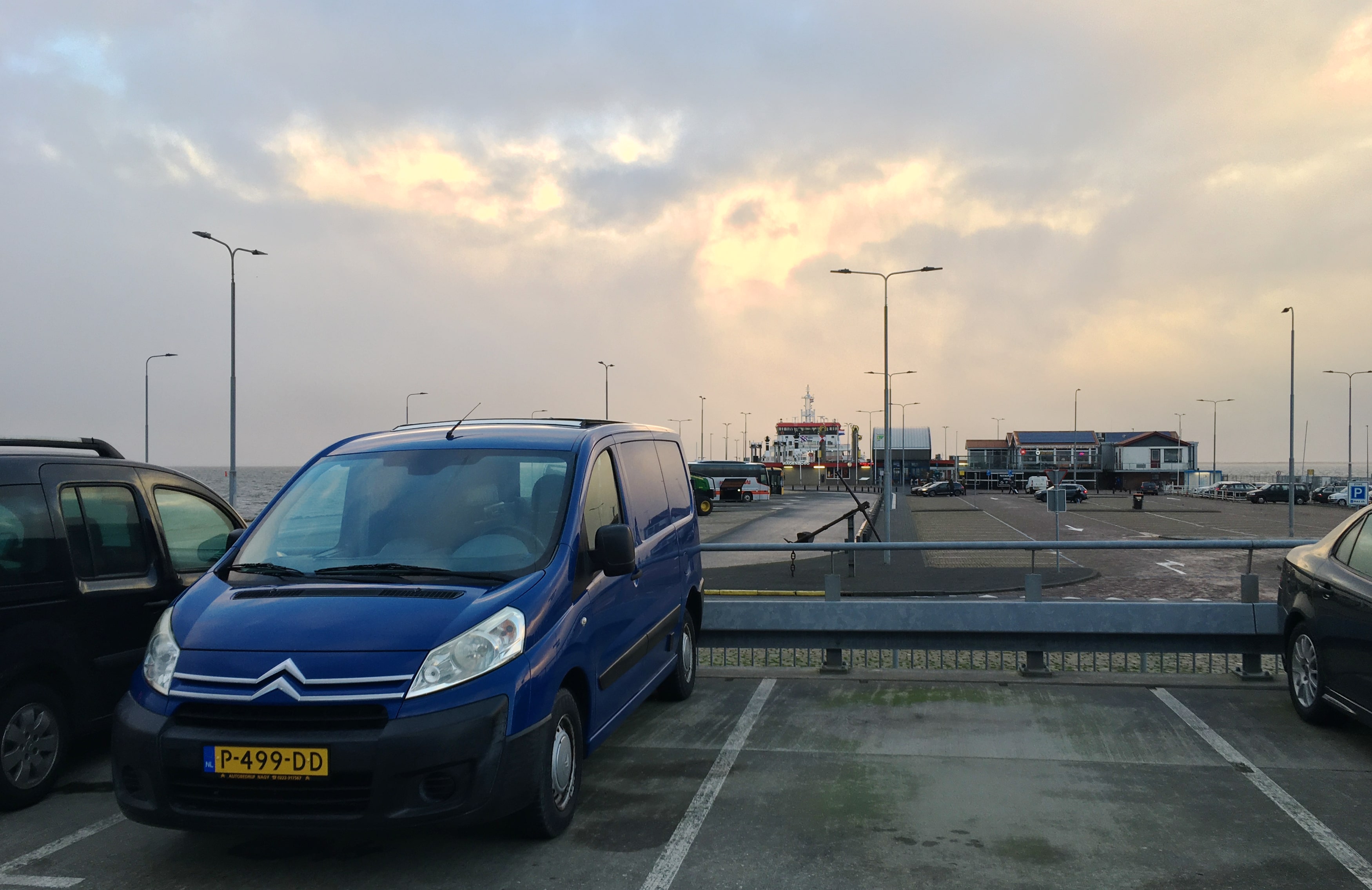 Parkeerplaats bij Veerboot Holwert. Op de voorgrond de Jumpie (Citroën bestelbus) op de achtergrond de steiger waar de boot net aankomt. De wolken gevulde lucht wordt wat warme kleuren gegeven door de ochtendzon.