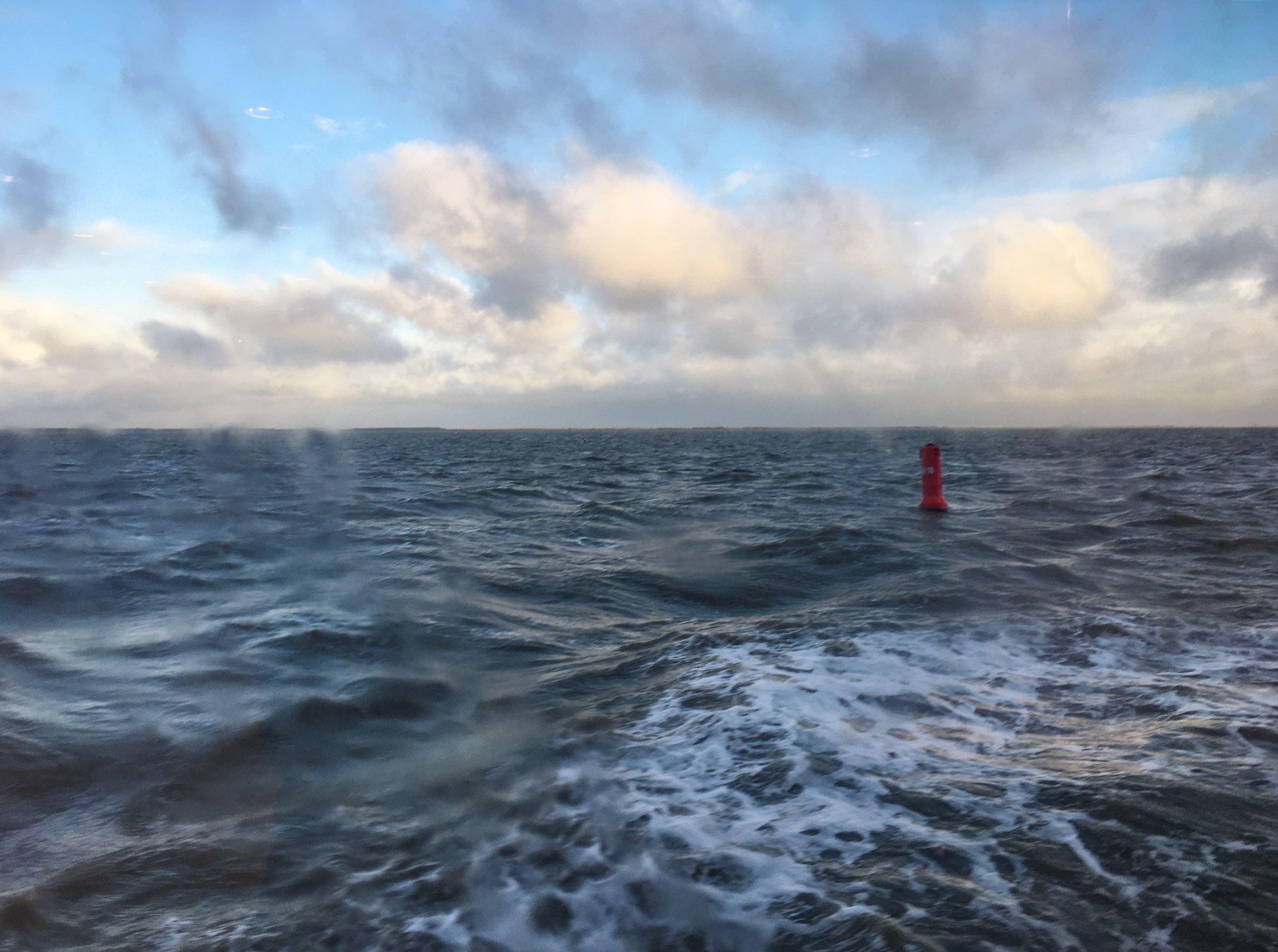 Waddenzee zover je kijken kan met stevige golven. Een rode boei en daarachter wat blauwe lucht en pastelkleurige wolken.