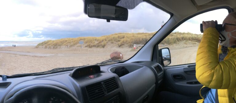 Vanuit de auto kijkend naar het Noordelijke stand van Texel. Je ziet links nog net wat zee en strand dan het begin van de duinen en achter de spijl van de auto kun je net een stukje vuurtoren zien. Els kijkt naar zee met de verrekijker.