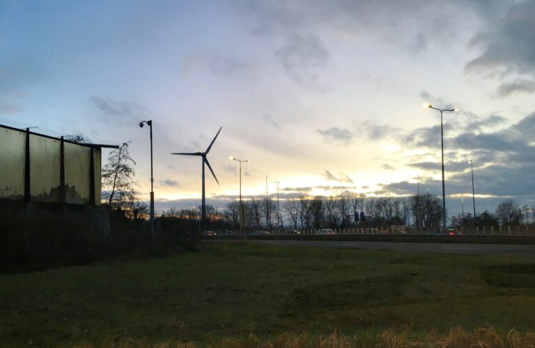 de zon is net onder en de foto laat een grasveld zien met daarachter een enkele windmolen en wat lantarenpalen die de snelweg erachter verlichten. In de verte wat inimini auotjes en bomen.