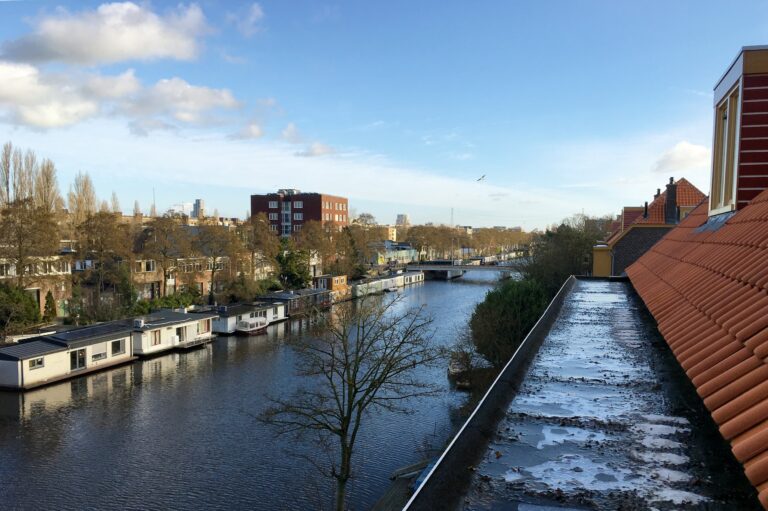 Uitzicht over het lozingskanaal vanaf het balkon op 3 hoog. Rechts op een platte strookje dak van ongeveer 50cm breed ligt ijs. Daarna begint het dak met rode dakpannen. Links op het kanaal woonboten en bomen. De zon schijnt en we hebben blauwe lucht met wat wolken.