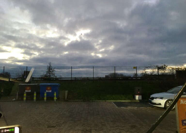 uitzicht vanuit autoraam bij tankstation. Je ziet recht de neus van een wit geparkeerde auto en links twee afvalcontainers. Daarachter een hek met het spoor van de metro. De lucht is bewolkt en de zon is net onder.