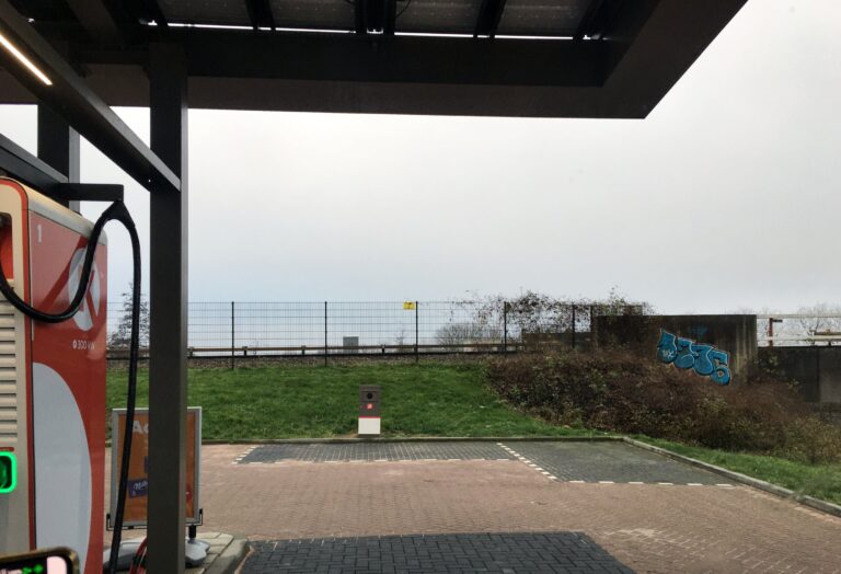 Terrein bij een tankstation. Links zie je de snellader,boven de overkapping. Wat parkeerplaatsen en rrachter een groene wal met een hek. Daar zit het spoor voor de metro verstopt. Het is een grijze dag.