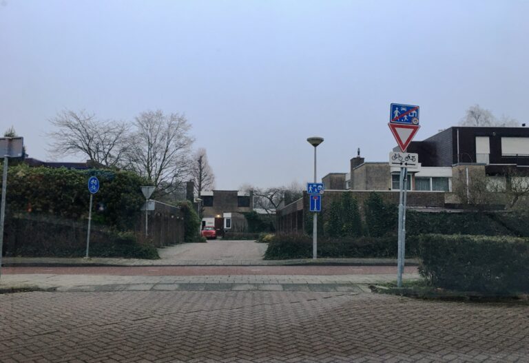 Inrit bij een woonwijk omringt door heggen. De huizen zijn semi vrijstaand, Twee hoog beetje als blokkendozen opgezet met wat verpringingen. Allemaal een plat dak. Geen hoge bomen.