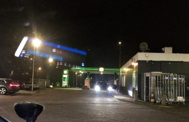 Uitzicht op een tankstation in het donker. Het is de neonverlichting en wat lantarenpalen die dit stukje wereld zichtbaar maken. Links wat geparkeerde auto's. Rechts de achterkant het gebouw met de winkel. Daar links van staat een auto geparkeerd waarvan de lichten recht op mij schijnen. Mijn telefoon spiegeld in mijn voorruit en zweeft wat in de lucht.