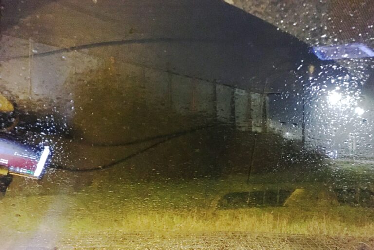 uitzicht vanuit mijn autoraam. Het is donker en het regent. Dus vol met druppels die het laat bijna niets zien wat voorbij het glas is. Je ziet vaag wat bosjes en een glaswand in een geel groen licht.