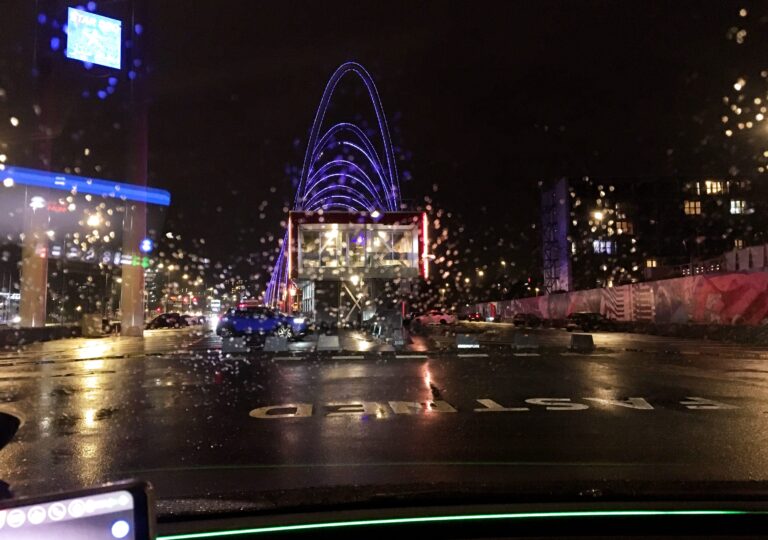 Uitzicht vanuit mijn autoraam van de foodstrip in Amsterdam Zuid-Oost. Het is donker en het regent, maar je ziet de neonverlichting In het midden waar de horeca zit. Links en rechts wat onduidelijke lichten van gebouwen die door de druppels verstrooit raken.