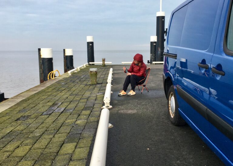 Het einde van de pier van Holwerd waar Els op een stoeltje zit in de zeer frisse licht. Rechts een deel van de bus. Op de achtergrond de Waddenzee en de horizon. Er zijn wat wolken in de lucht. De zee is kalm.