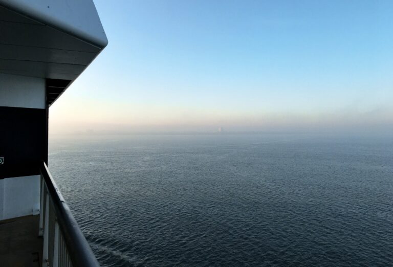 Uitzicht vanaf de Waddenzee op Den Helder die net zichtbaar wordt door de mist. De zon komt net op en geeft een oranje gloed. De lucht is blauw, de zee is kalm. 