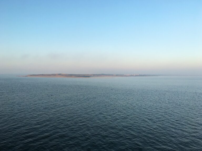 Uitzicht vanaf de Waddenzee op de Mokbaai van Texel. De zon komt net op en de lucht is wolkloos maar er hangt een ochtendmist die bedekt en een roze gloed geeft.