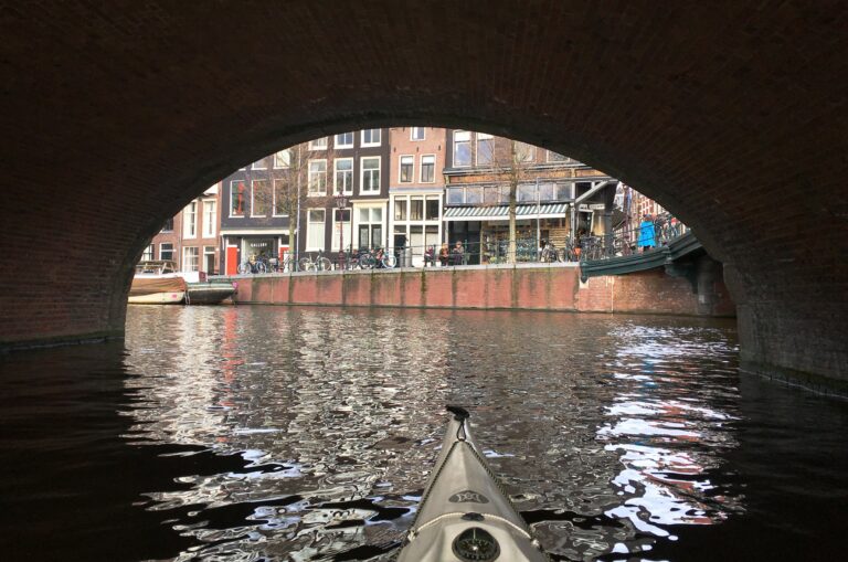 Onder een stenen brug met de karakteristieke boog. Aan het eind zie je delen van de kade en de typische 19e-eeuwse bouw.
