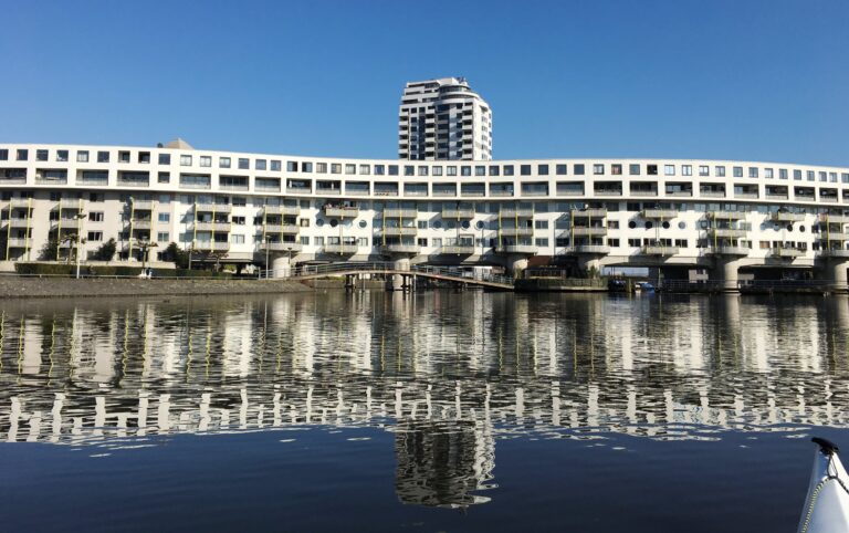 Een wit gebouw over de hele breedte van de foto met de spiegeling ervan in het water. Door de kleine rimpelingen is het vervaagt maar brengt een mooi symmetrisch en contrastrijk beeld met boven en onder blauw van de lucht.