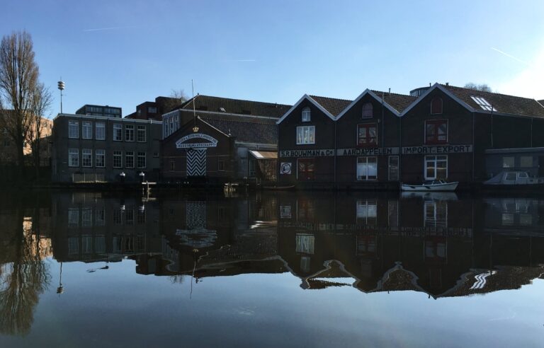 Oude pakhuizen en langs het water. De lucht is blauw, het water als een spiegel. Waardoor het silhouet van de gebouwen herhaald wordt in het water