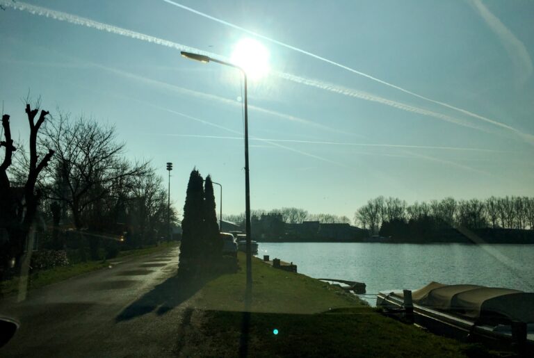 uitzicht vanuit de auto met rechts water en links wat bomen. De foto is donker omdat ik tegen de zon in fotografeer die als witte vlek in de blauwe lucht hangt
