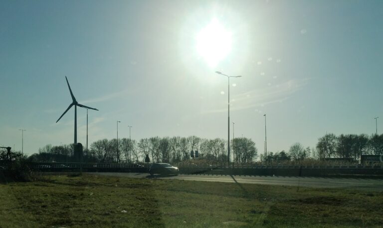 Uitzicht vanuit mijn auto op de snelweg. Er rijden wat auto's op en er is een windmolen te zien. De foto is donker omdat de zon fel schijnt en ik er teginin fotografeer. 