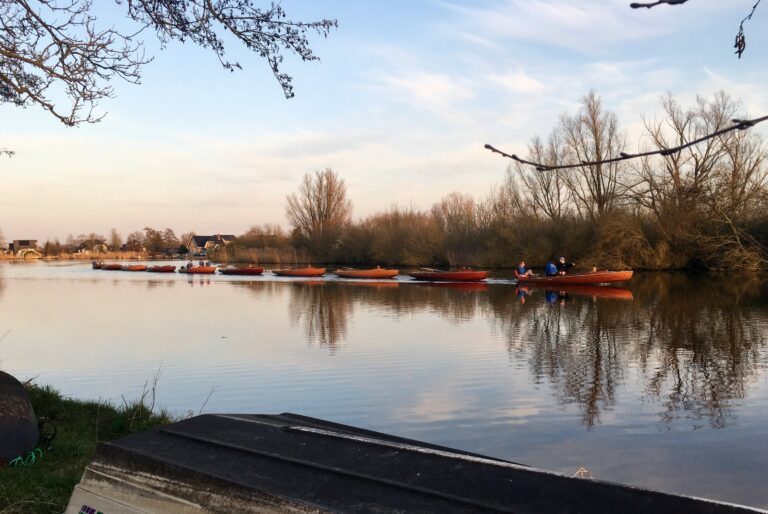 Op het stille water van de rivier, terwijl de zon ondergaat en warm licht verspreid, varen 10 houten zeilbootjes met hun masten naar beneden en aan elkaar verbonden terug naar de haven. 