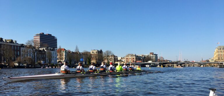 Acht mans roeiboot met stuurman/vrouw en aanjager/coach om de terugweg na de race van 8 kilometer naar de Amstel in Amsterdam. Op de achtergrond de westoever van de Amstel die afsteekt op een blauwe lucht. Het water is kalm.