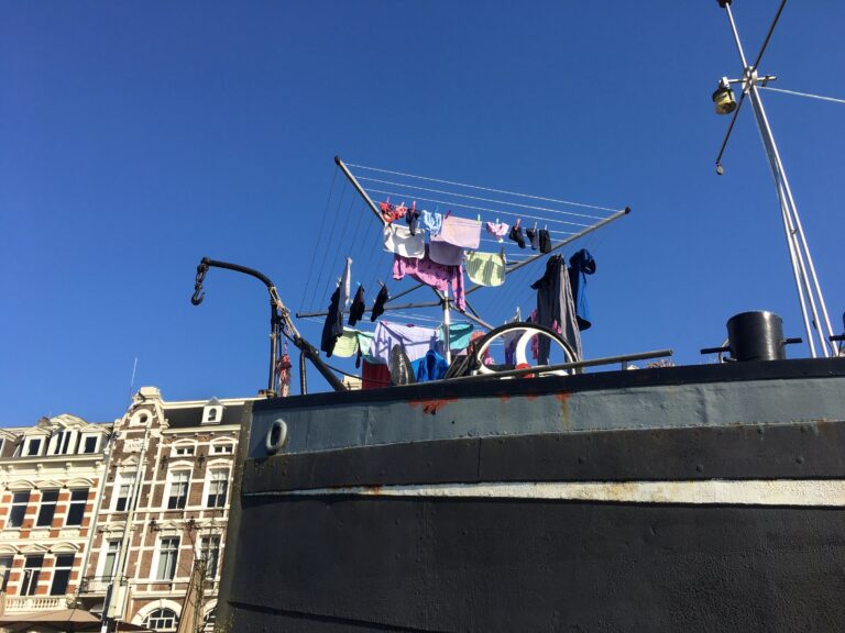 Boeg van een schip met daarop een droogrek met was. links nog wat klassieke gevels van huizen langs de Amstel in Amstedam