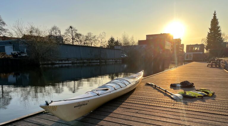 Mijn witte kajak op de stijger bij KV Zeeburg. Rechts op de steiger de peddel, het zwemvest en met spatzeil. Links het water en de zon is aan het ondergaan schrijnt een warm geel licht.