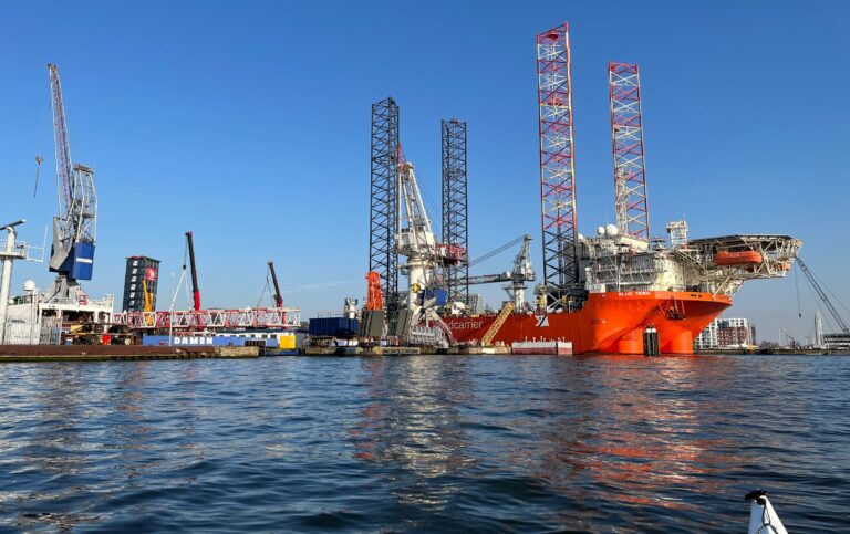 foto vanaf het water van de windcarrier Fred. Olsen. Een schip met een helikopter platform en vier grote pijlers die het naar de bodem kan brengen zodat het boven het water uit kan staan. Nu is dat ook het geval al is het minimaal. Links daarvoor de drogdoks van scheepswerf DAMEN met ander schepen en kranen.