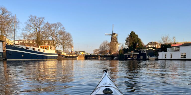 Panorama van de Oostenburgergracht in Amsterdam. In het midden zie je de Dagenraad brug en daarachter molen De Gooyer. De lucht is blauwe en het water kalm.