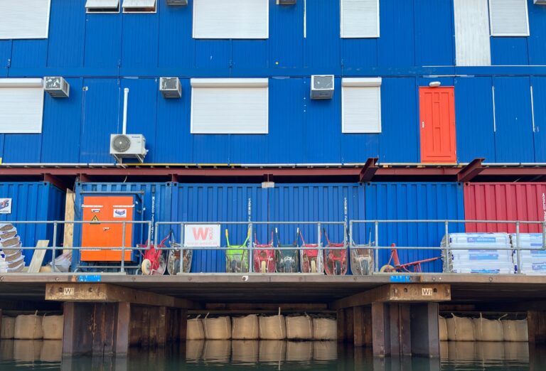 Blauwe bouwketen en containers op pale i het water met daarvoor een omgang waar allemaal zwaar gebruikte kruiwagens op een rijtje staan tegen de container.
