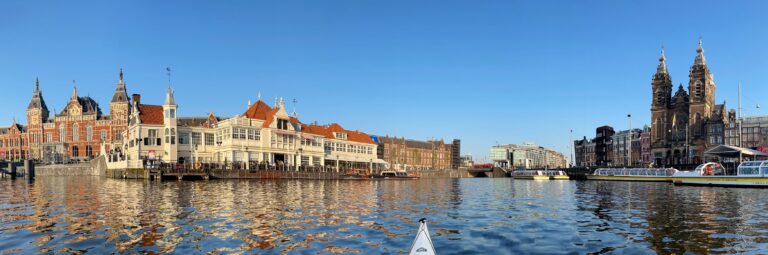 Panorama vanaf het water van het Centraal Station in Amsterdam, het Noord-Zuidhollands koffiehuis en de Basiliek van de Heilige Nicolaas.