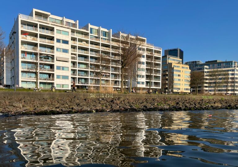 Vanaf het water is de oever te zien van basaltblokken met daarboven weer een betonnen rand. Daarachter flatgebouwen van 8 verdiepingen. Op de betonnen rand van de oever staat grafiti "SHELL IS EVIL"