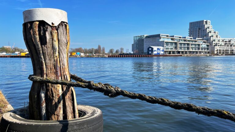 Een meerpaal met een stak gespannen touw met een lus er omheen. De paal is oud en verweerd nt als het touw. Op de achtergrond water met aan de andere oever moderne witte flatgebouwen van 6 tot 10 hoog.
