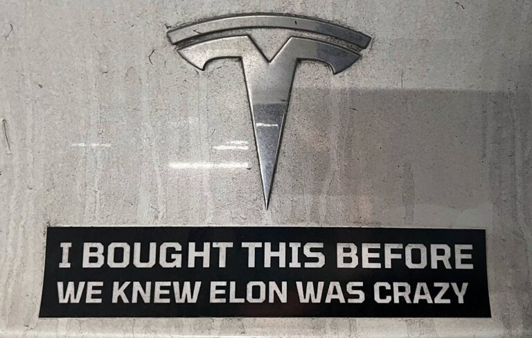 Tesla logo op de achterkant van een auto met daaronder een zwarte stikker met witte tekst in blokletters "I bought this before we knew Elon was crazy"