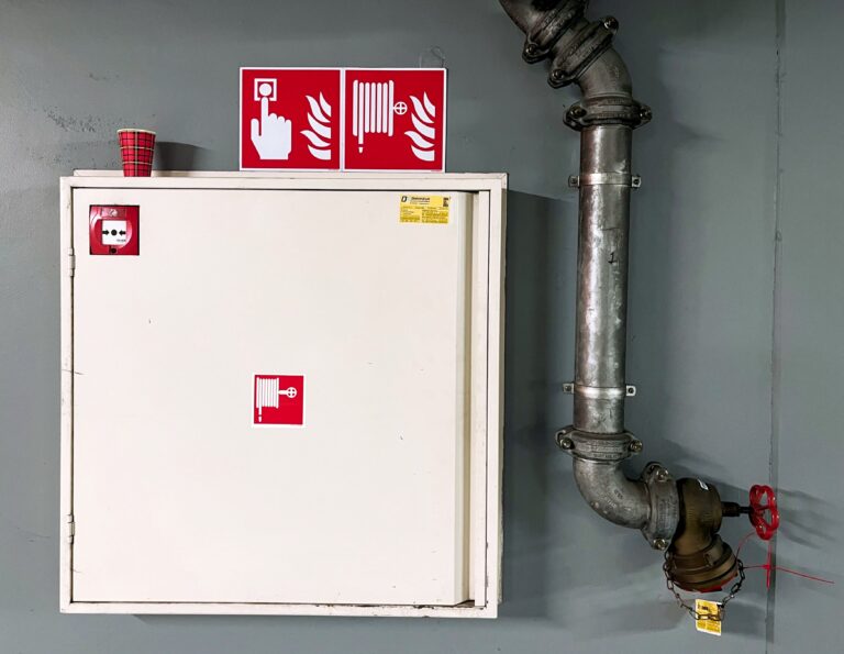 Witte brandslangkast op grijze muur. Voorzien van rode bestickering en bordjes zodat iedereen het weet.  Rechts de grote vuistdikke pijp waar de slang op aangesloten kan worden. Naast die rode bordjes op de kast staat links een papieren rood koffiekopje met ruitmotief.