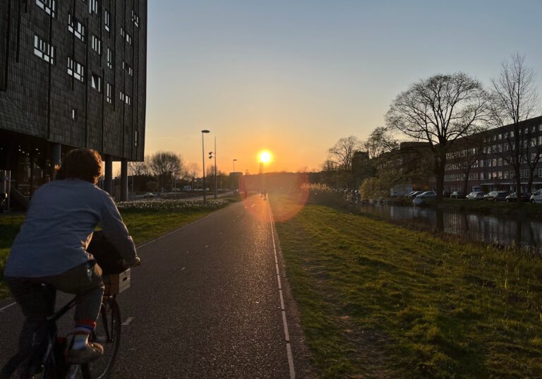 Een recht fietspad op een dijkje met het zonlicht aan het einde. Wodt net ingehaald door een fietser en de aura van de rond fietst ook ng een groepje mensen. We gaan naar het licht. Rechts water en daarachter weer gebouwen van 4 verdiepingen. Links een groot flatgebouw dicht op de dijk.