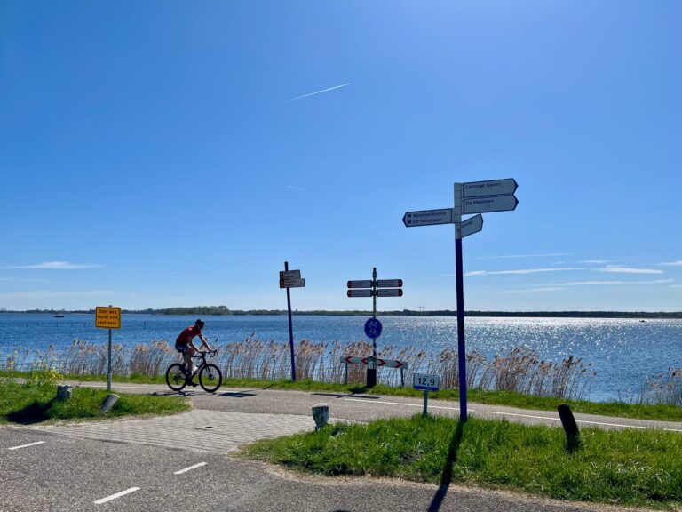 Uitzicht over het Gooimeer op een stralende zonnige dag met een blauwe hemel en glinsterend blauw water. Op de voorgrond de oever met een strookje gras en een racefietser en allemaal bordjes naar de diverse bestemmingen.