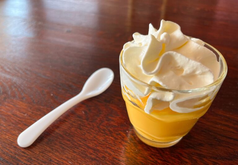 Een glaasje advocaat met een toefje slagroom. Ernaast een wit lepeltje. En dit alles op een glimmend houten tafelblad.