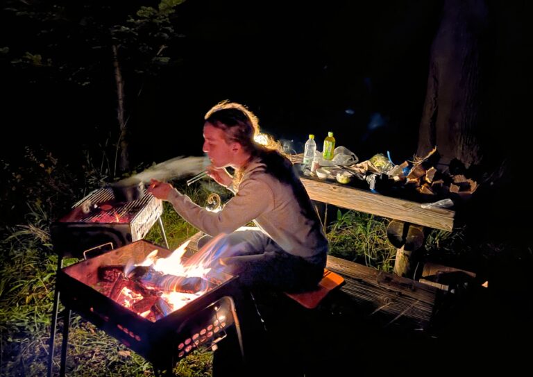 Zoon in zijn element met links van hem een vuurtje, voor hem het gloeiende houtskool en rechts al het eten op de picknickbank. In het pikkedonker op gras.
