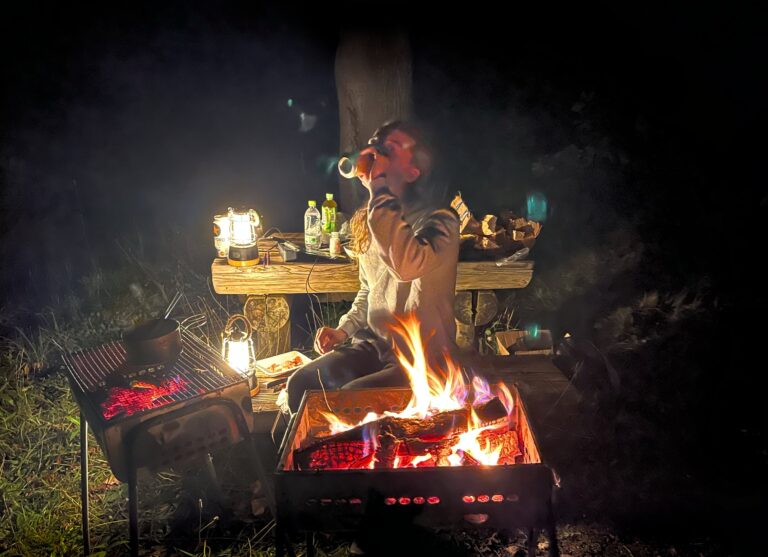 Zoon in zijn element met links van hem een vuurtje, voor hem het gloeiende houtskool en rechts al het eten op de picknickbank. In het pikkedonker op gras.